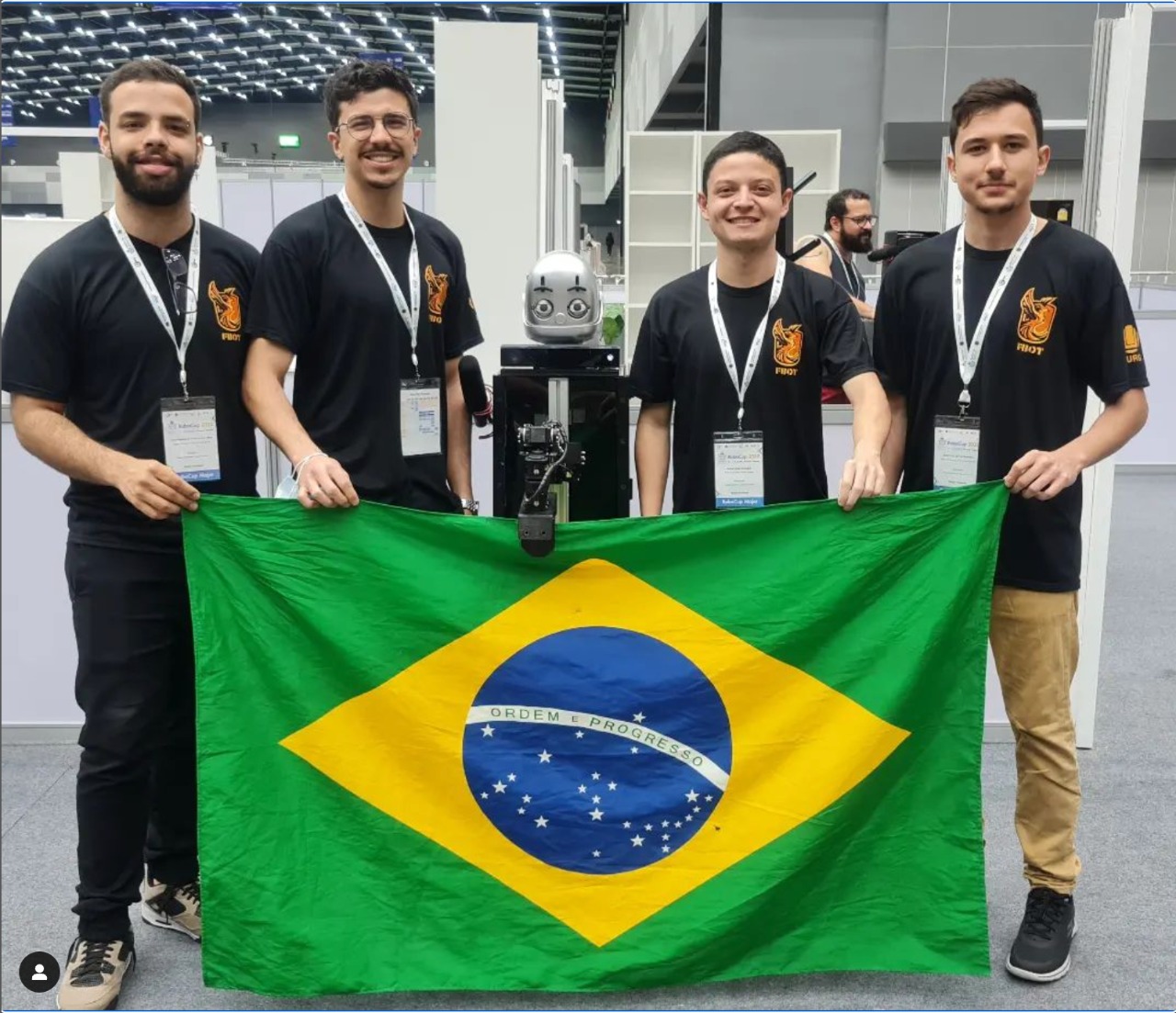 quatro alunos vestindo camiseta preta ao centro um robo. ambos seguram a bandeira do Brasil