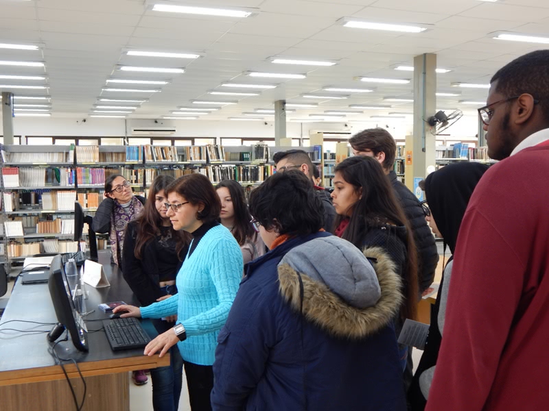 alunos aparecem em pé, ao redor do monitor do balcão de atendimento da biblioteca. todos observam atentamente a explicação da bibliotecária